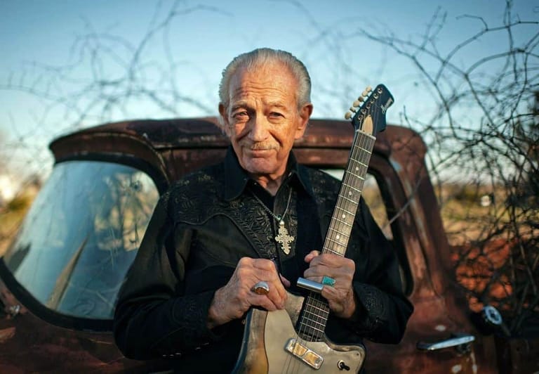 Older man in black shirt holding guitar stands before rusted truck in arid landscape.