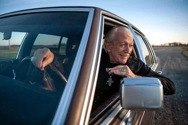 Man leaning out of car window holding a small white container in an open landscape.