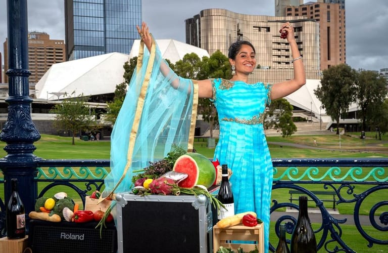 Woman in bright blue dress holds patterned fabric above fresh produce at an outdoor Adelaide venue with city skyline behind.