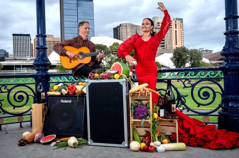 Two musicians perform with guitar and vocals on a decorated bridge, with city skyline and festive garland backdrop.
