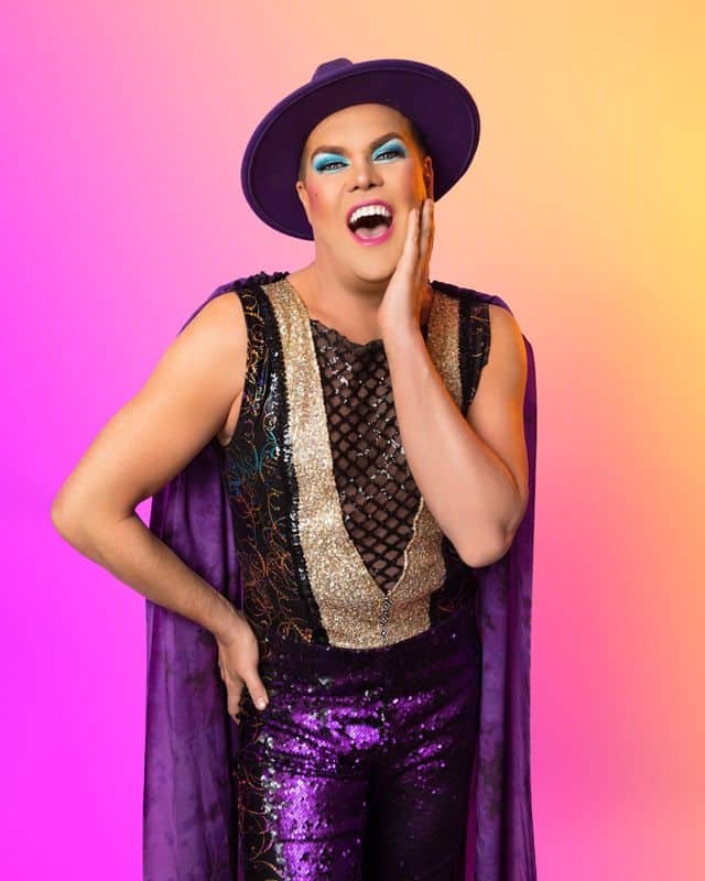 Performer in sequined purple pants and black vest with purple cape against a vibrant pink and yellow gradient background.