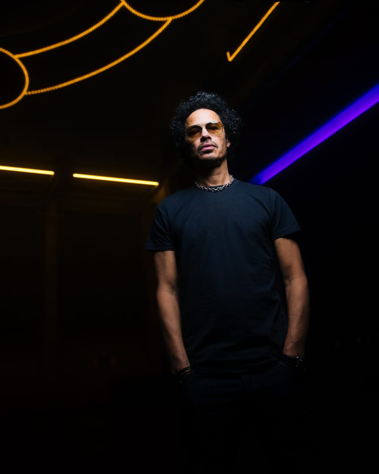 Man in black t-shirt and sunglasses stands against neon yellow and blue stage lighting.
