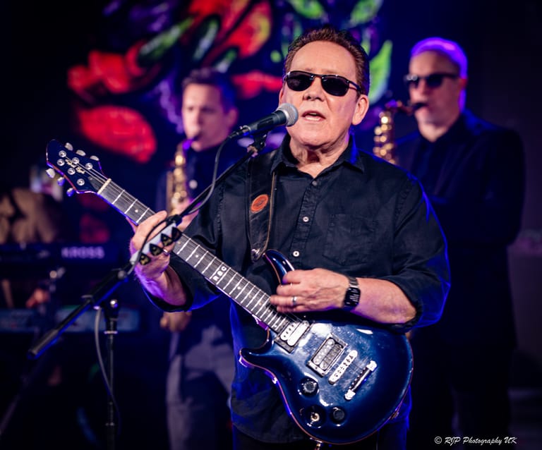 Guitarist in sunglasses performing with electric guitar on stage under coloured lights, backed by band members.
