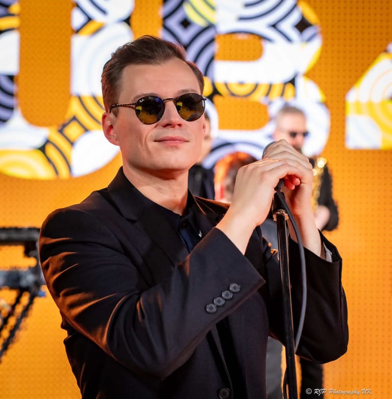 Performer in dark suit and sunglasses singing into microphone against bright yellow branded backdrop.