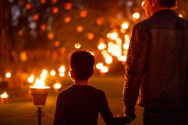 Two figures hold hands gazing at glowing lanterns strung overhead in warm orange light.