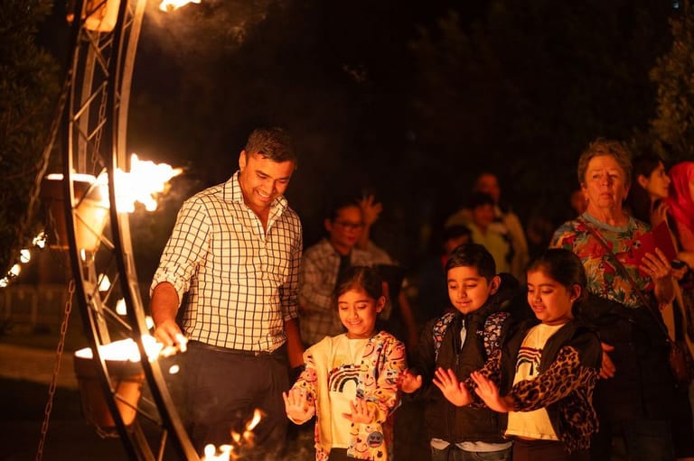 Man in checked shirt leads children in performance around illuminated fire pit at night.