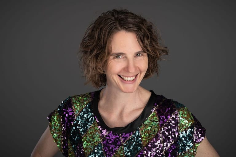 Person wearing a sequined top with purple, green and silver panels smiles against a grey background.