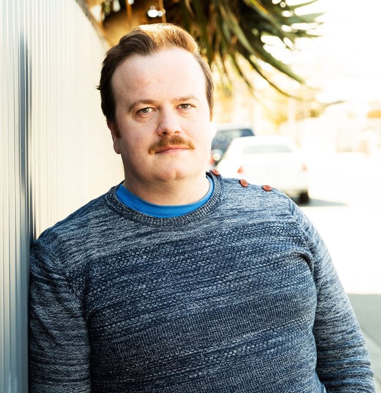 Man with styled hair and moustache wearing a blue-grey knit jumper, leaning against a white wall with tropical plants visible behind.