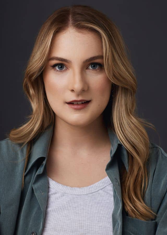 Woman with long honey-blonde wavy hair wearing a grey jacket over a white top, against a dark neutral background.