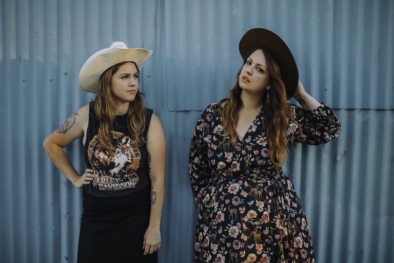 Two women in cowboy hats stand against a corrugated metal wall, one wearing a graphic tank top and the other a floral dress.