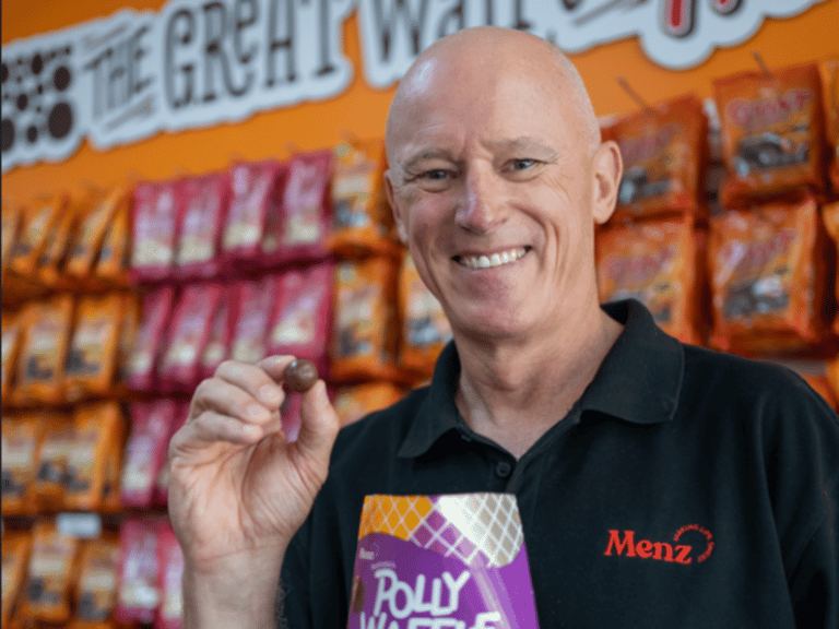Man in black polo shirt holding Polly Wafer product with fist raised, standing before colourful merchandise display.