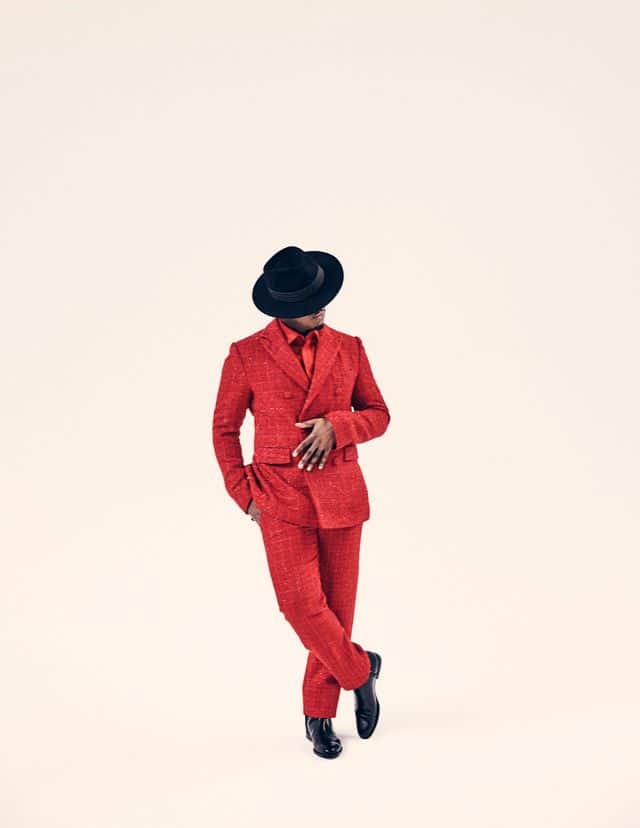 Person in a bright red suit and black fedora hat posed against a pale background.