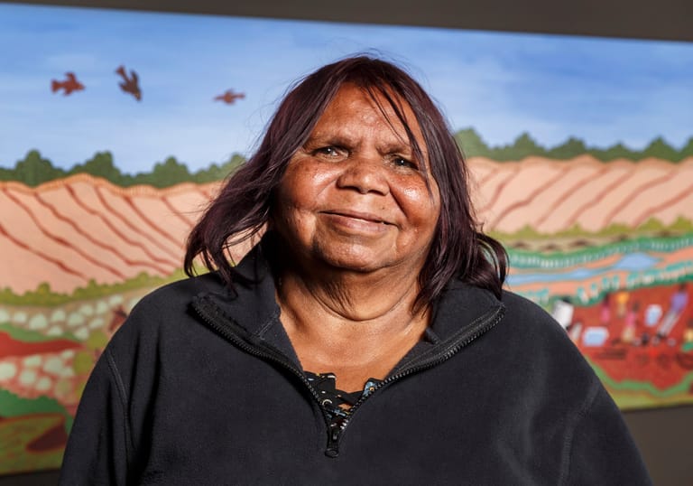 Woman with dark hair in black hoodie smiling before agricultural landscape artwork with red earth and blue sky.