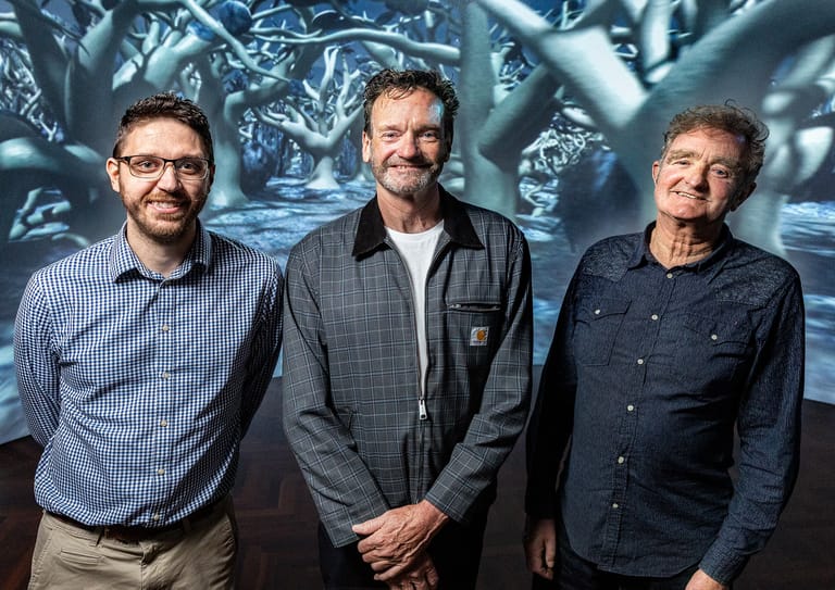 Three men stand in front of an ornate metallic relief sculpture backdrop.