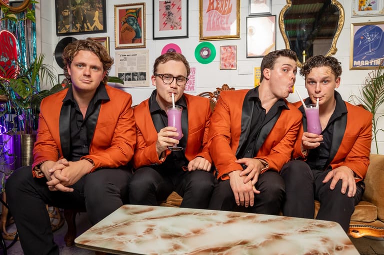 Four musicians in matching orange jackets sit together on a couch in a colourful, poster-covered room.