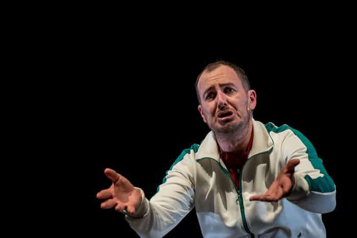 Man in cream and teal jacket gestures expressively while speaking against a black background.