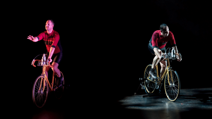 Two performers on bicycles lit by pink and blue stage lights against a black background.