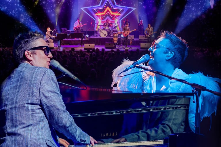 Two musicians perform at a grand piano on stage under purple lighting, with a neon star backdrop and band visible behind them.
