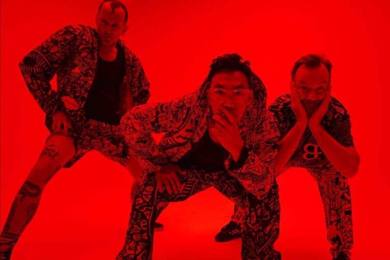 Three performers in patterned outfits pose dynamically against a bright red backdrop.