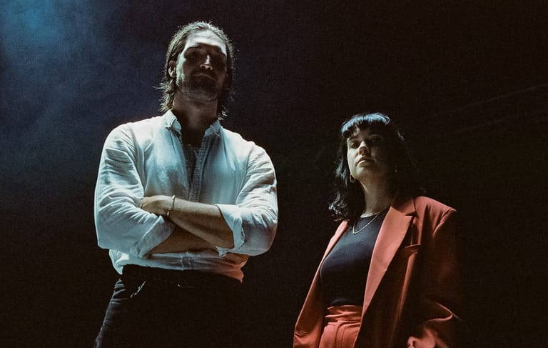 Two performers on stage in dramatic blue and warm lighting, one in a light blue shirt with arms crossed, the other in a coral jacket.