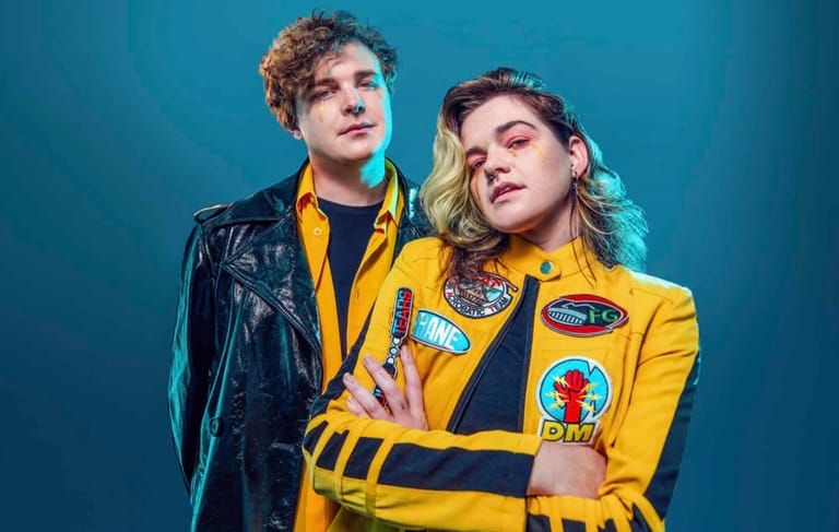 Two performers in bold yellow and black jackets pose together against a blue backdrop.