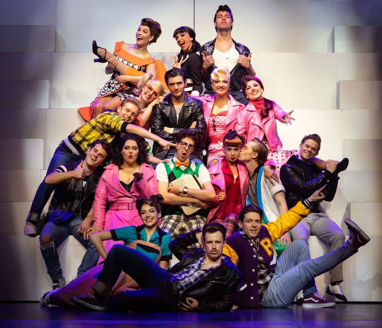 Large ensemble cast in vibrant costumes posed dynamically on stage under warm lighting.