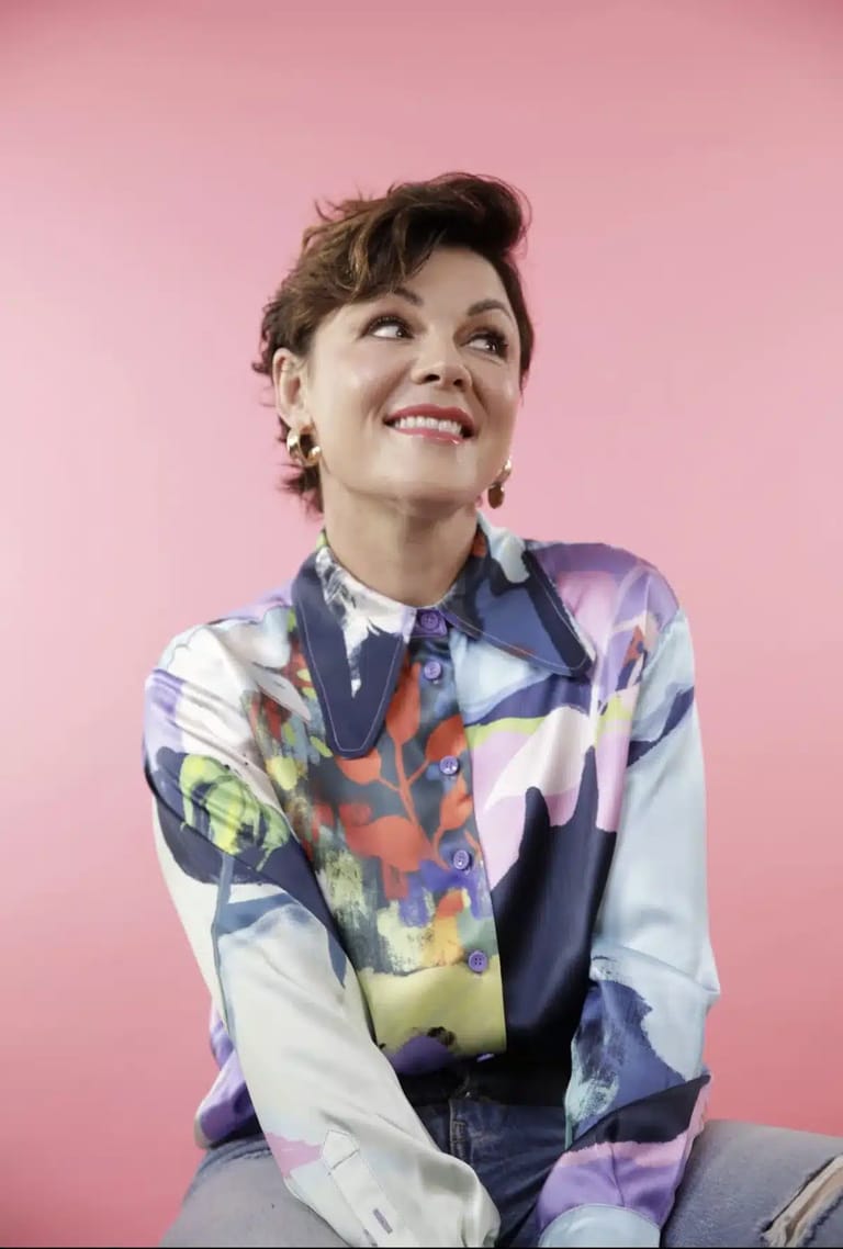 Person with short dark hair wearing a colourful abstract patterned shirt against a pink background, smiling upward.