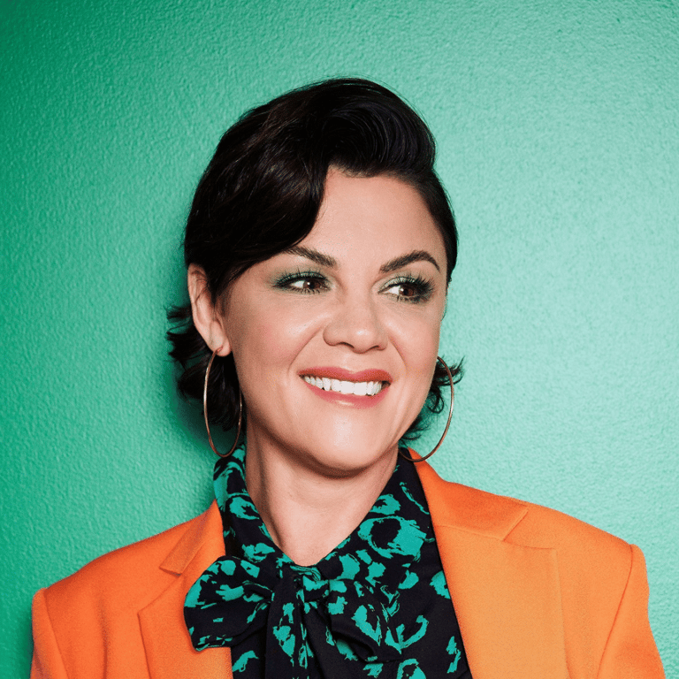 Woman with dark hair smiling against a turquoise backdrop, wearing a bright orange blazer over a patterned shirt.