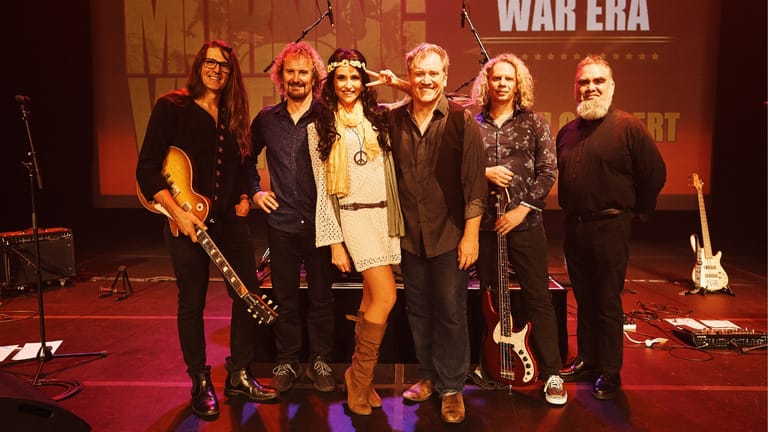 Six musicians stand on a stage lit in warm orange and red tones, with "WAR ERA" visible on the backdrop behind them.