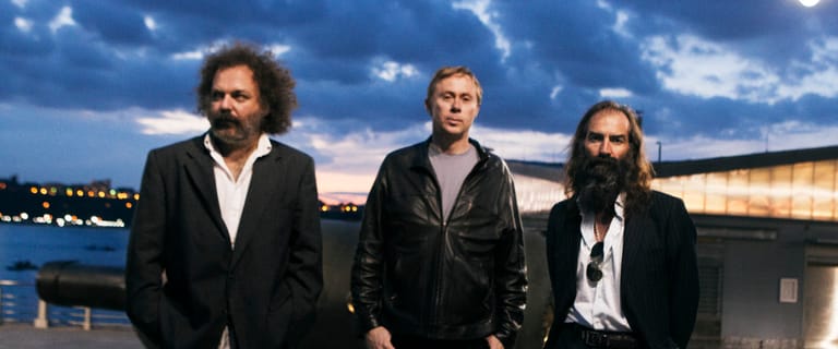 Three men in dark jackets stand against a twilight waterfront backdrop with illuminated buildings.