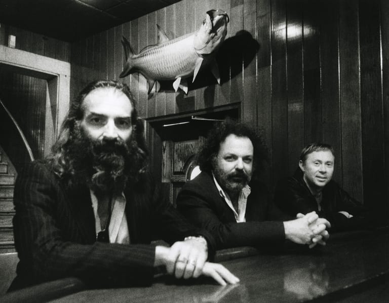 Three bearded men in dark suits sit at a table with a taxidermied animal head mounted on the wall behind them.