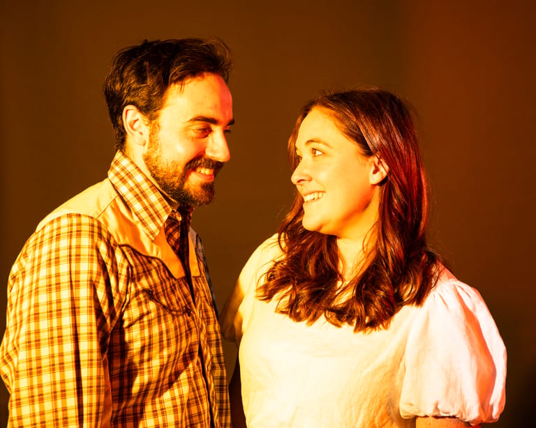 Two performers face each other on stage under warm amber lighting during a theatrical scene.