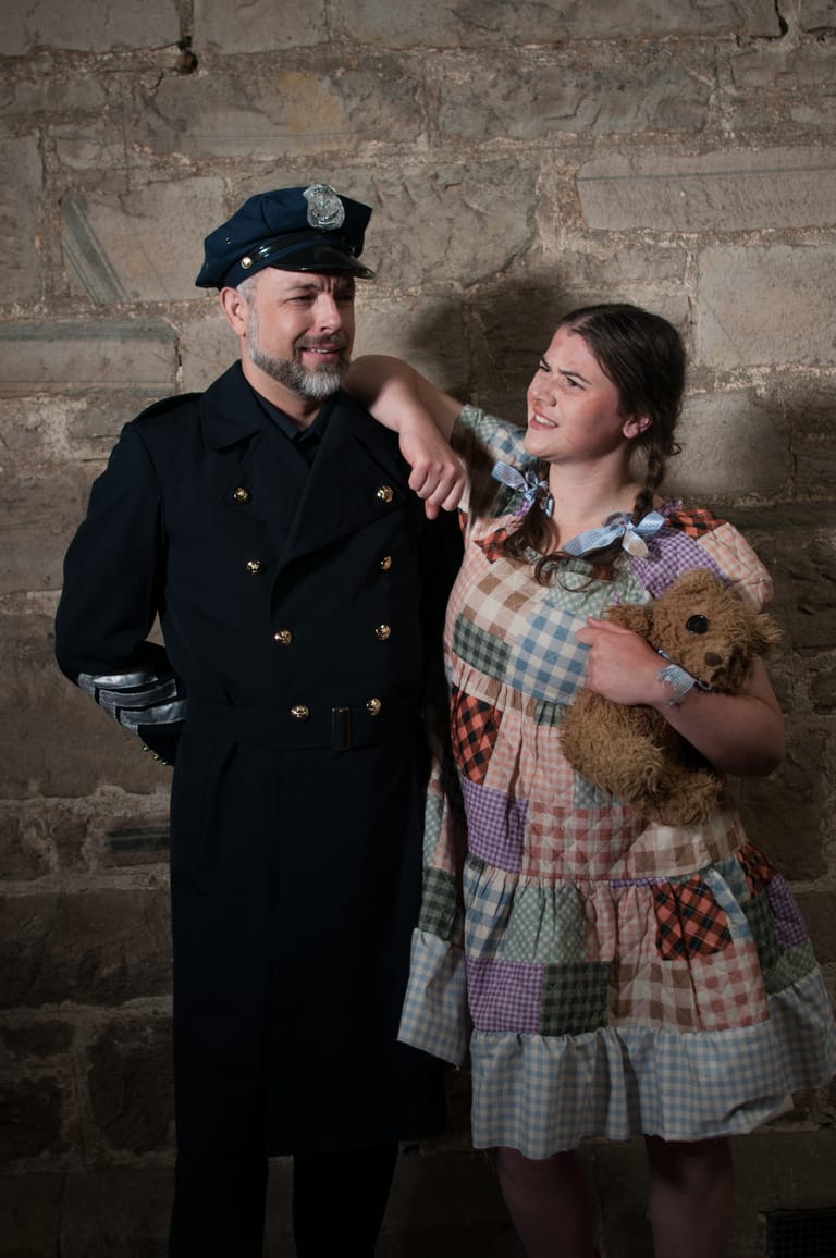 Man in naval uniform and woman in patchwork dress hold a teddy bear against a weathered brick wall.