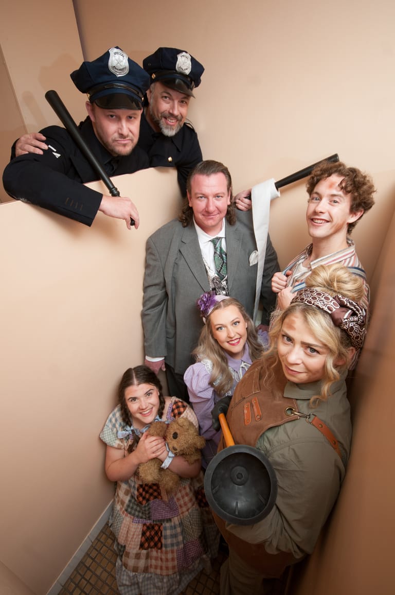 Six cast members in period costumes and props pose together overhead shot against a neutral backdrop.