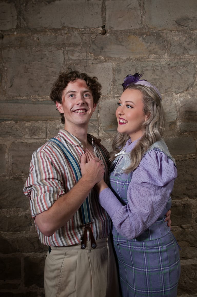 Two performers in vintage-style clothing smile whilst posing against a weathered stone wall backdrop.