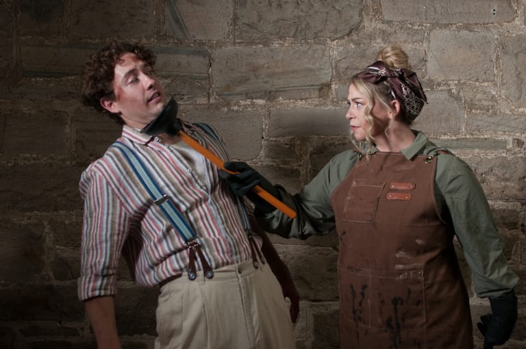 Two performers in period costume stand against a stone wall during a theatrical scene.