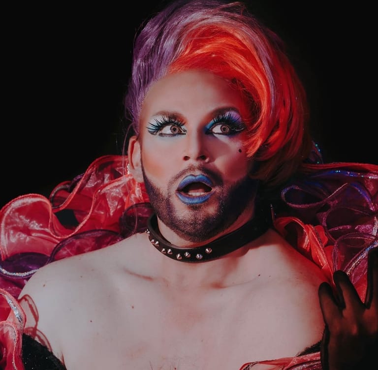 Performer with striking red and purple hair, bold makeup and studded choker against a black background.