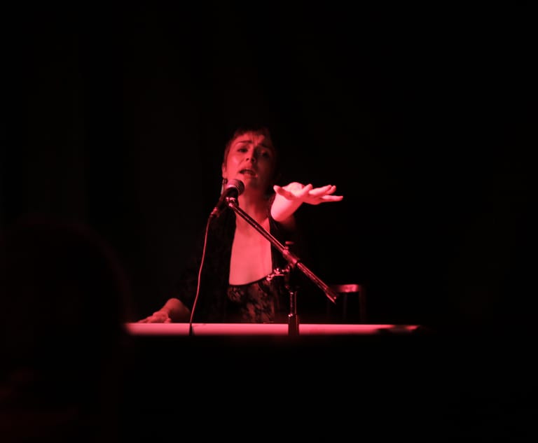 Performer at microphone on stage, illuminated by red spotlight against black background.