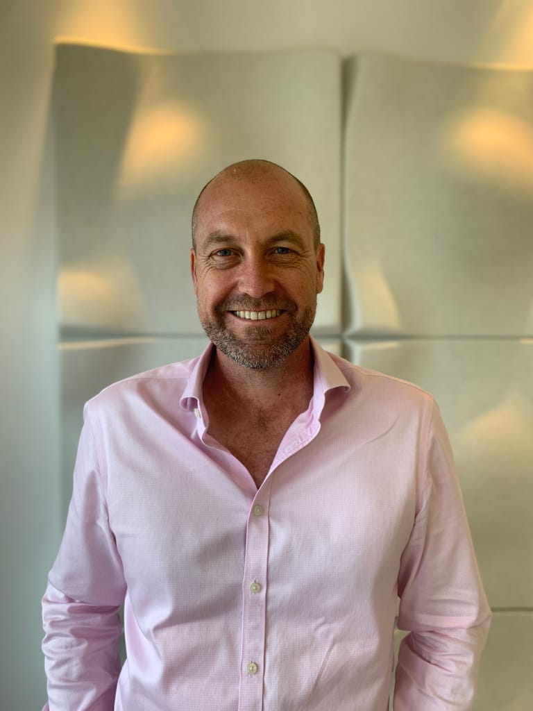 Man in pink shirt smiling at camera against geometric wall backdrop.