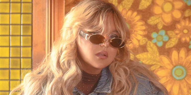 Blonde woman wearing oval sunglasses and denim jacket against a warm-toned, patterned interior backdrop.