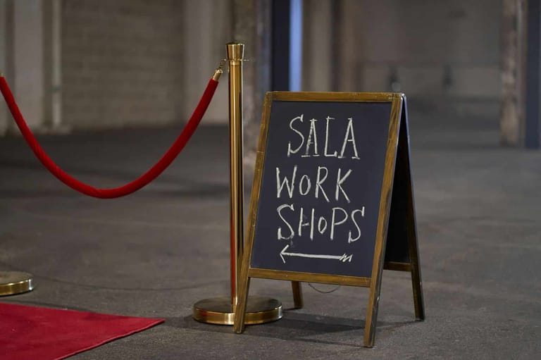 Chalk board sign reading "SALA WORK SHOPS" stands on floor near red velvet rope and brass stanchion.