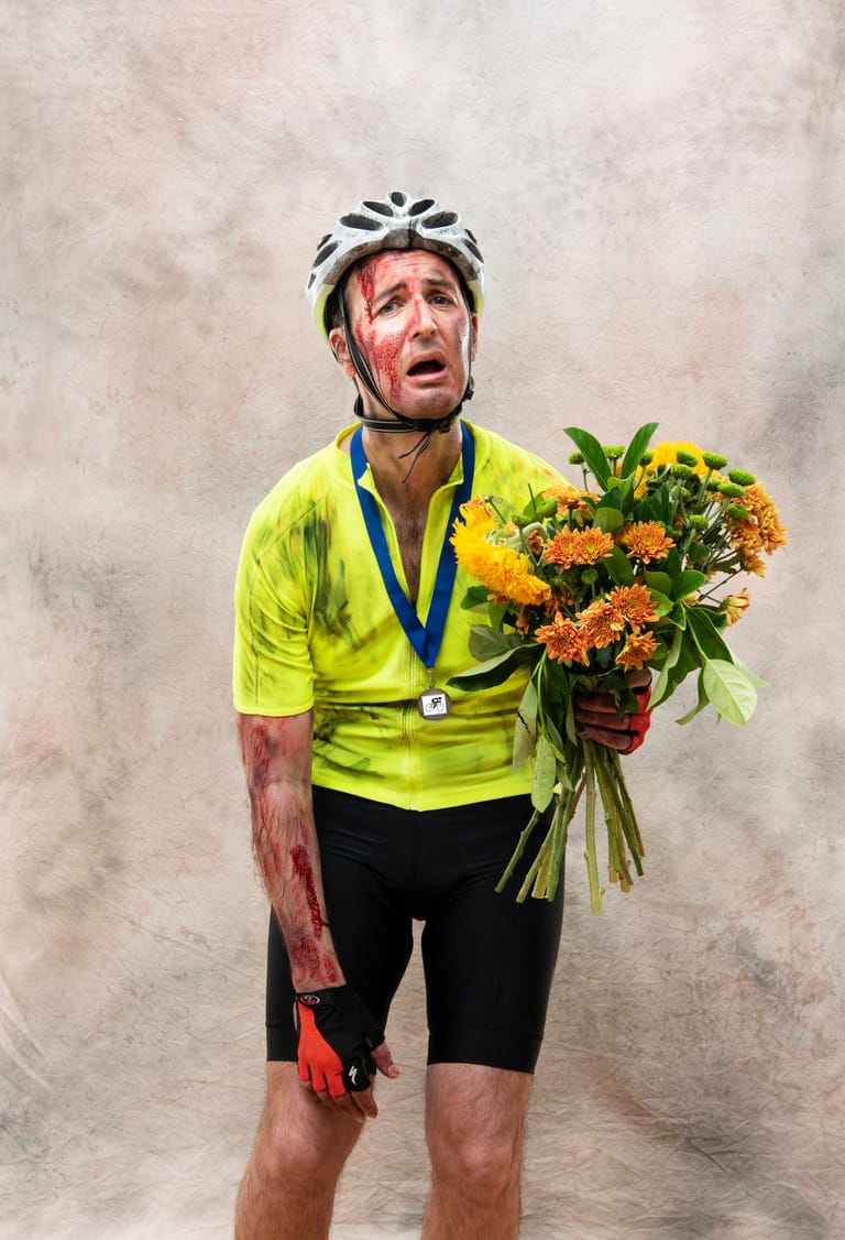 Cyclist in neon yellow jersey and helmet holding orange flowers and medal against grey backdrop.