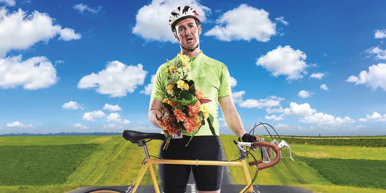 Cyclist in high-visibility gear holds a bouquet while standing with bike on a rural road under blue sky.