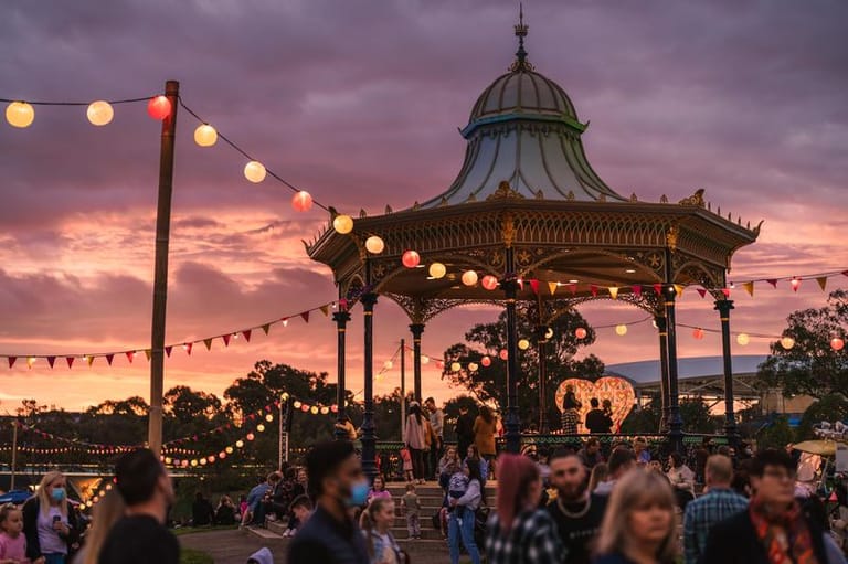Ornate domed gazebo illuminated by string lights as crowds gather during a purple sunset.