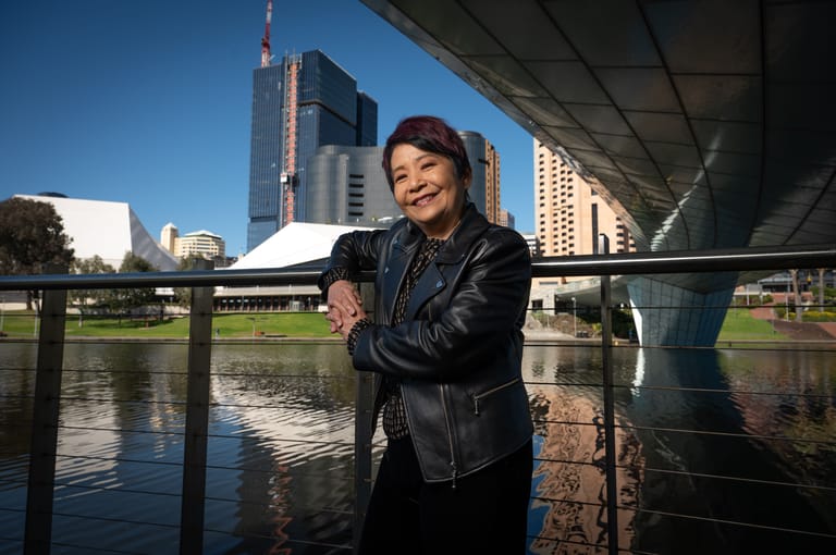Woman in black leather jacket smiles at camera beside Adelaide riverbank with city skyline and bridge behind.