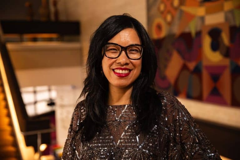 Woman with long black hair and glasses wearing a sparkly grey top, smiling in an indoor venue setting.