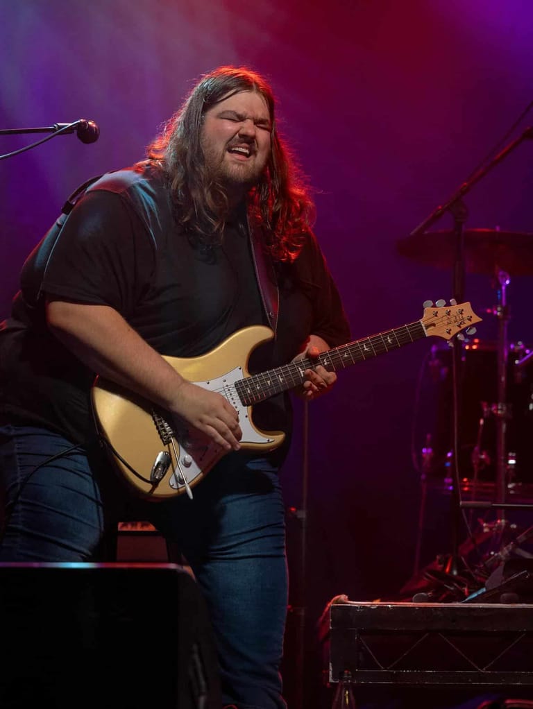 Guitarist with long dark hair performing on stage with electric guitar under purple and red stage lighting.