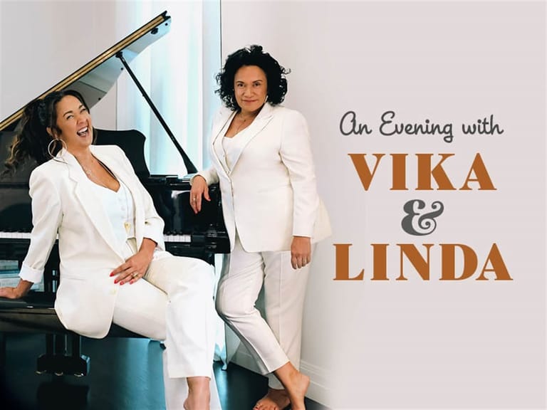 Two musicians in white outfits pose by a grand piano for "An Evening with Vika & Linda" promotional image.