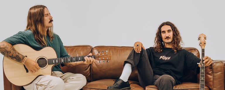 Two musicians sit on a brown leather couch with guitars, against a grey background.