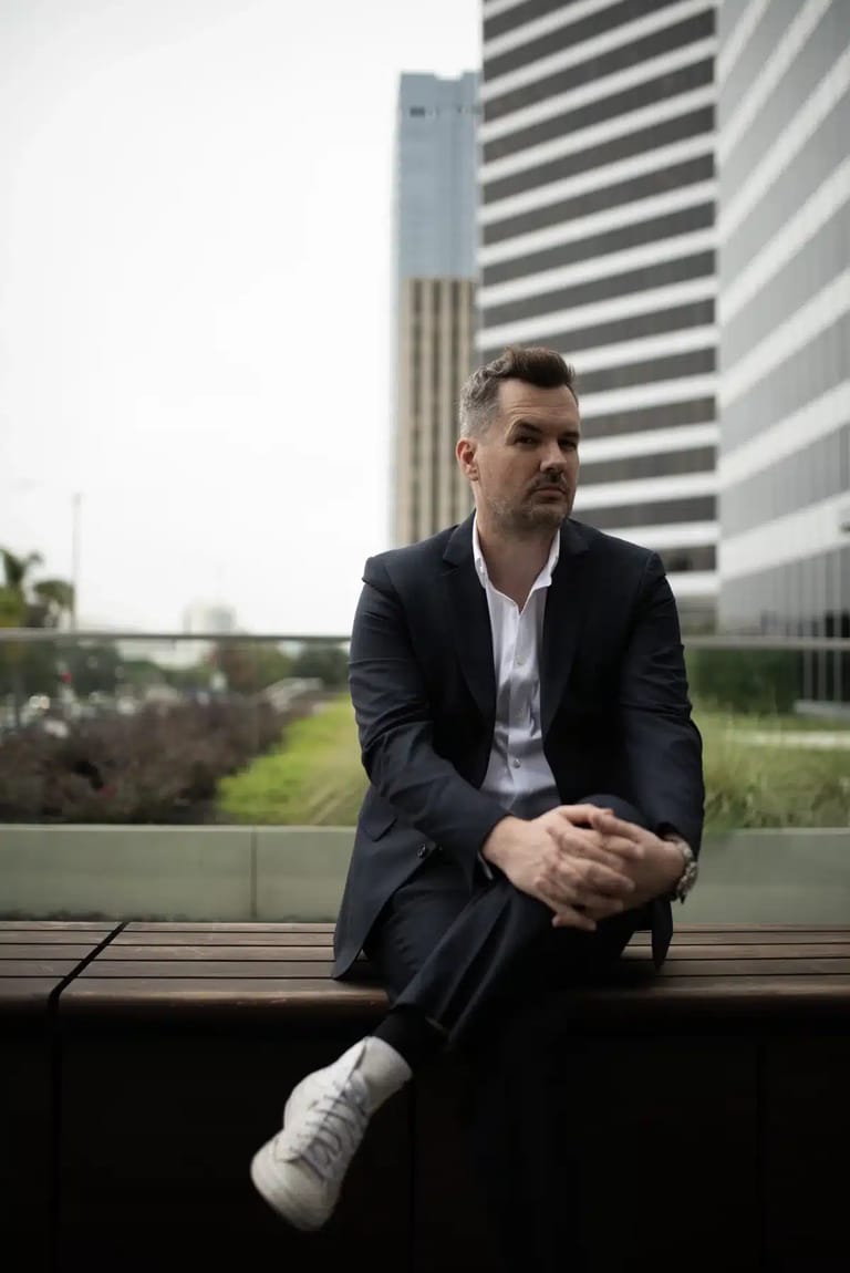 Man in dark blazer seated on a bench with modern high-rise buildings in the background.